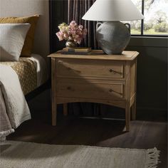 a nightstand with flowers on it next to a bed in a room that has a large window