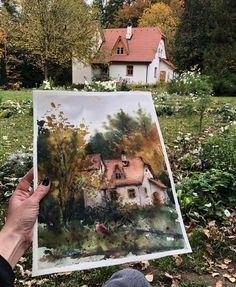 a person holding up a painting in front of a house with autumn leaves on the ground