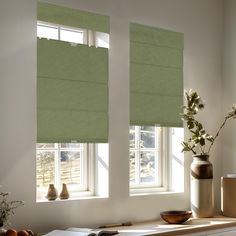 two windows with green roman shades in a white room next to vases and flowers