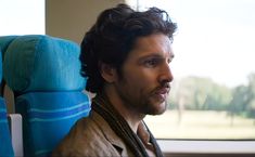 a man with curly hair sitting on a train looking out the window at something in the distance