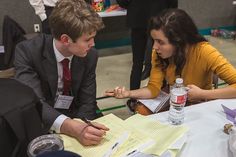 two people sitting at a table with papers and pens in front of them, talking to each other