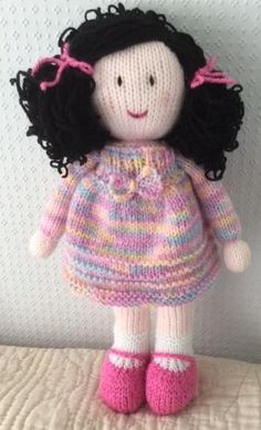 a knitted doll with black hair wearing a pink dress and pink shoes on a bed