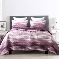 a bed covered in a purple and white comforter next to a night stand with two nightstands