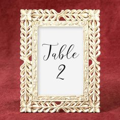 a gold frame with the word table 2 in black ink on it, sitting on a red velvet surface