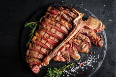 two hands holding a knife and fork over a steak on a black plate with sprigs of rosemary