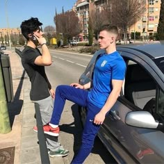 two people standing next to a car talking on their cell phones