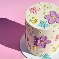 a white cake with purple and blue flowers on it sitting on a plate next to a cupcake