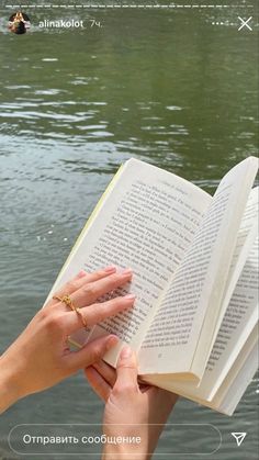 a woman is holding an open book in her hands near the water, with one hand on top of it