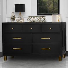 a black dresser with gold handles and drawers in front of a window, next to a lamp