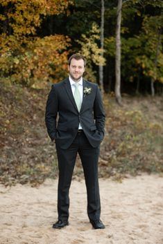 a man in a black suit and green tie standing on the sand with trees in the background