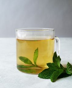 a glass cup filled with green tea next to some mint leaves on a white surface