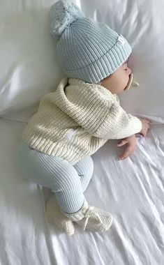 a baby laying on top of a white bed wearing a blue hat and sweater with pom poms