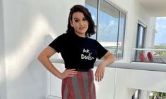 a woman standing in front of a window with her hands on her hips wearing a black shirt and striped skirt