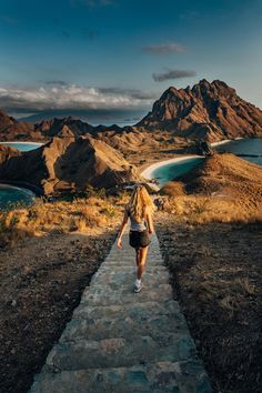 a woman walking down a stone path towards the ocean and mountains with blue water in the background