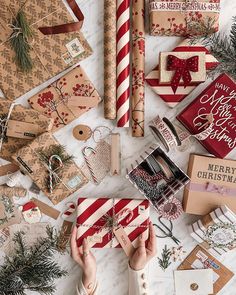 someone wrapping christmas presents on a table with holiday cards and other items around them, including wrapped gifts