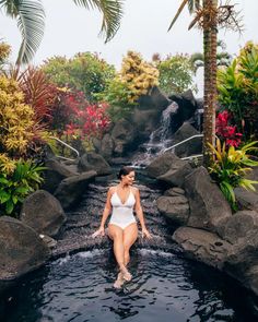 a woman in a white swimsuit sitting on rocks near a waterfall and palm trees