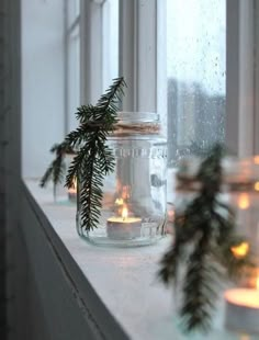 two jars with candles are sitting on a window sill next to evergreen branches and pine cones