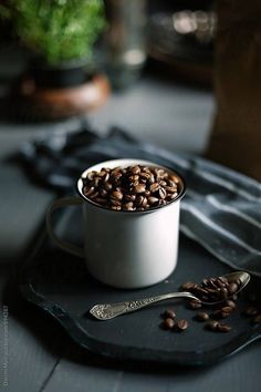 a white cup filled with coffee beans on top of a black tray next to a spoon