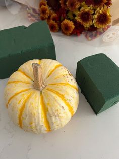 there is a small pumpkin next to two blocks of green foam on the table with flowers in the background