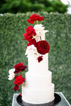 a three tiered white cake with red and white flowers on the top is surrounded by greenery