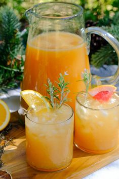 two glasses filled with orange juice and garnished with rosemary sit on a cutting board