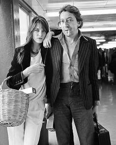 a man and woman standing next to each other in front of a wall holding luggage