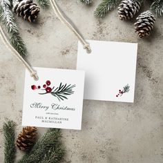 two christmas cards sitting next to pine cones and evergreen branches with red berries on them