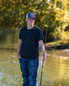 a man standing in the water while holding a fishing pole and wearing a hat with his hands behind him