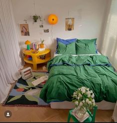 a bed with green sheets and pillows next to a yellow table in a room that has white curtains