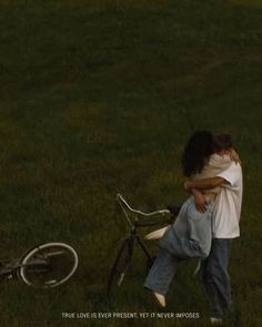 two people hugging each other in front of a bike on the grass with an empty field behind them