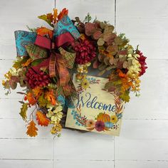 a welcome wreath with autumn leaves and ribbons on a white brick wall behind it is a sign that says welcome