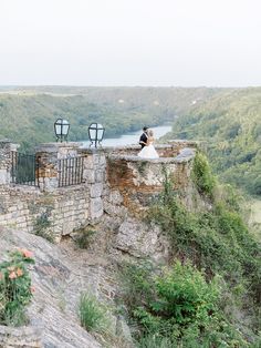 a bride and groom are sitting on the edge of a cliff looking out over a valley