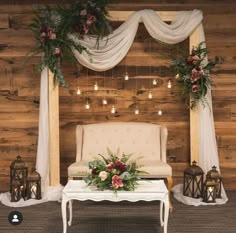 a chair and table with flowers on it in front of a wooden wall decorated with hanging lights