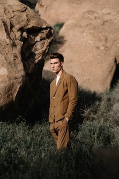 a man standing in the grass next to large rocks and bushes with his hands in his pockets