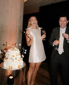 two people standing next to each other holding wine glasses in front of a wedding cake