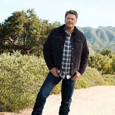 a man standing on top of a dirt road in front of trees and mountains with his hands in his pockets