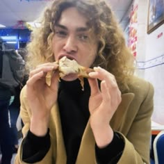 a man with curly hair eating food in a room full of other people and looking at the camera