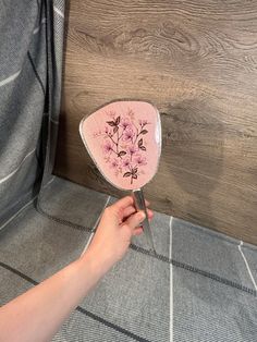 a hand holding a pink flower shaped mirror on top of a wooden floor next to a wall