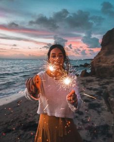 a woman holding sparklers in her hands on the beach at sunset with water and clouds behind her