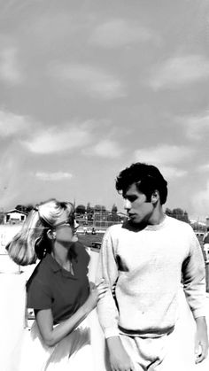 black and white photograph of man and woman walking on beach with boats in the background