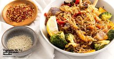 a white bowl filled with noodles, broccoli and other vegetables next to a wooden spoon