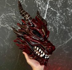 a hand holding a red dragon mask on top of a gray surface with cracks in it