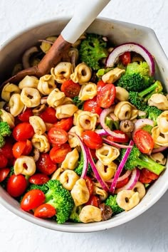 a white bowl filled with pasta and veggies next to a wooden utensil