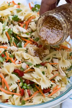 a person pouring dressing onto a pasta salad in a glass bowl with carrots, celery, and noodles