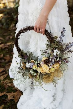 a woman in a white dress holding a basket filled with flowers and greenery on her wedding day