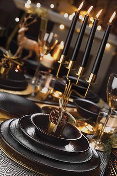 a table set with black plates and gold place settings, candles and deer figurines