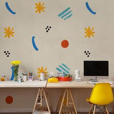 a desk with two chairs and a computer monitor on it in front of a colorful wall