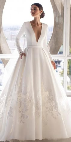 a woman standing in front of a window wearing a white wedding dress with long sleeves