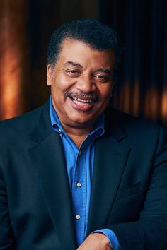 a smiling man in a black suit and blue shirt with his arms crossed, looking at the camera