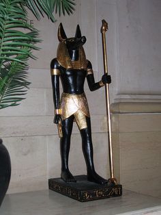 a statue of an egyptian god holding a staff in front of a potted plant